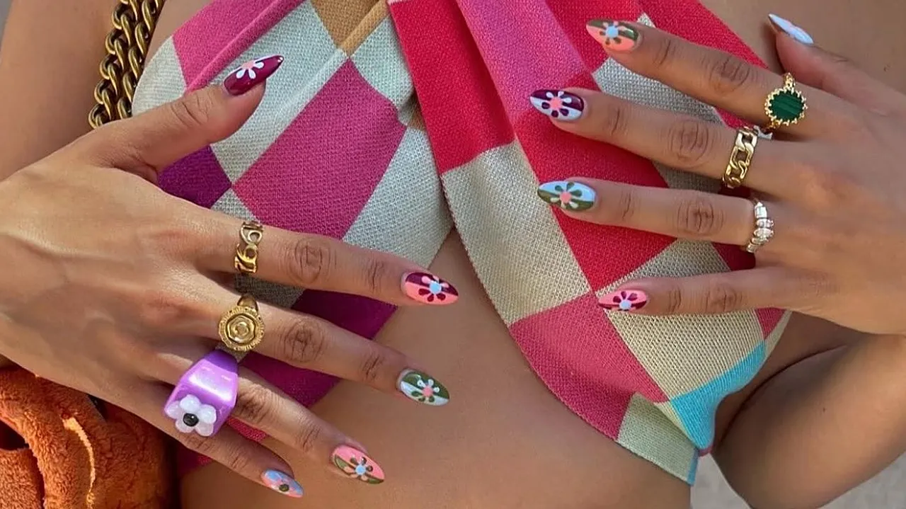 Närbild på händer med färgglad handmålad floral nail art och guldringar.